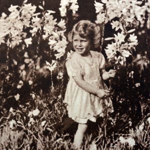 Child stands holding a flower, surrounded by tall blooming lilies in a garden.