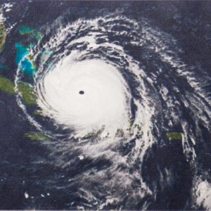 Satellite hurricane spins over Caribbean islands, dense clouds swirling dramatically, surrounded by dark ocean waters.