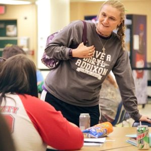 A person stands talking to seated individuals in a cafeteria setting, with snacks and drinks on the table. Text on shirt: 