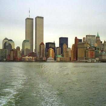 Skyscrapers stand tall, reflecting cityscape in water, under an overcast sky with a boat's wake in the foreground.