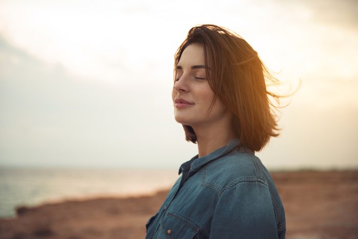 Great pleasure. Charming calm young woman is standing near sea with closed eyes and expressing delight. She is posing against wonderful sunset while enjoying last rays of the sun