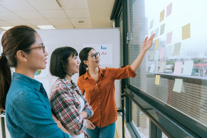smiling beautiful woman manager showing idea for her designer team and stick many memo paper on office glass window wall during meeting.