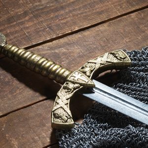 A decorative sword with a gold hilt rests on chainmail atop a wooden surface, showcasing intricate medieval designs.