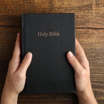 Hands hold a black book titled "Holy Bible" on a wooden table.