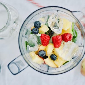 Blender holds ice, berries, and peach slices ready for blending; surrounded by scattered blueberries, raspberries, and a whole apple on a white surface.