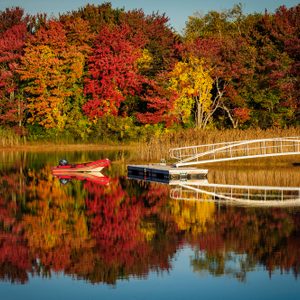 A red boat rests on a calm lake reflecting vibrant autumn trees and a curved white dock in serene, colorful surroundings.