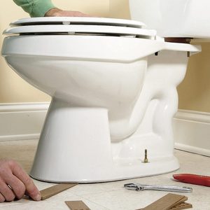 Toilet being installed; tools and cardboard on tiled bathroom floor, hand adjusting alignment.