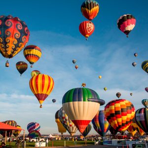 Hot air balloons ascend vibrantly against a clear blue sky, above a grassy field with spectators and colorful tents.