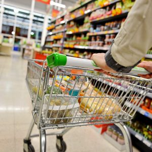 A person pushes a shopping cart filled with groceries through a brightly lit supermarket aisle.
