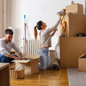 Couple packs cardboard boxes, organizing items in a bright, modern living space with a wooden floor and nearby kitchen.