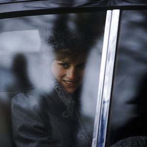 Smiling person sits inside a car, wearing a fur hat, visible through a frosted window in a wintry environment.