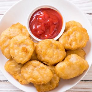Chicken nuggets sit on a white plate with a small bowl of ketchup, placed on a wooden table.