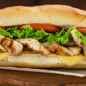A sub sandwich filled with grilled chicken, lettuce, tomato, and cheese, resting on parchment paper on a wooden surface.
