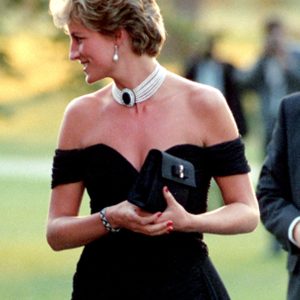 A person in elegant attire holds a clutch, standing outdoors with blurred people in the background, exuding a formal event atmosphere.