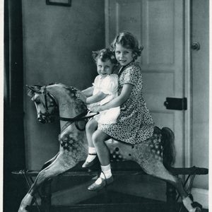 Two children sit on a wooden rocking horse, smiling, in a room with a closed door and framed picture on the wall.