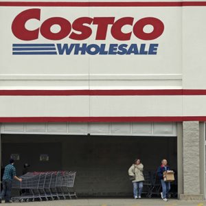 Shoppers exit Costco, pushing carts and carrying boxes, outside a large retail entrance with prominent 
