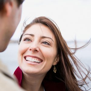 A smiling woman looks upward, with hair blowing in the wind, beach background visible.