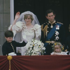 Bride waves on a balcony, groom standing in uniform beside her; surrounded by children in formal attire, red draped railing in foreground.