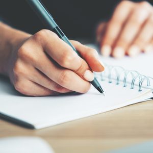 A hand writes with a pen in a spiral notebook on a wooden desk.