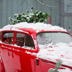 A red car; covered in snow; parked outside with a tree on the roof and heart shape on windshield.