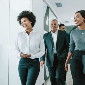 People walk and smile in a bright office corridor, holding folders and dressed in business attire.