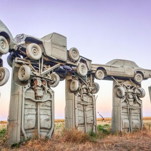 Cars are stacked vertically, resembling a stonehenge formation, set in an open grassy field with a clear sky background.