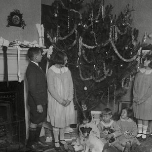 Children admire a decorated Christmas tree beside a fireplace with stockings and presents, a dog and two kittens sit nearby on the floor.