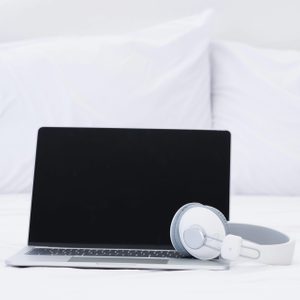 A closed laptop sits beside white headphones on a bed, with white pillows in the background.