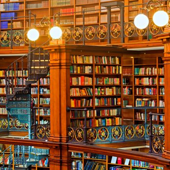 Books fill shelves in a multi-level library, featuring ornate railings and a spiral staircase, under warm lighting.