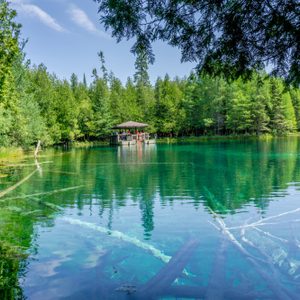 A wooden pavilion stands over turquoise water surrounded by lush green trees under a clear blue sky.