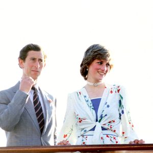 Two people stand smiling, one raising a fist slightly. They are on a boat, with a clear sky behind them.
