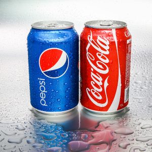 Pepsi and Coca-Cola cans stand side-by-side on a wet surface. The background is shiny and covered with water droplets.