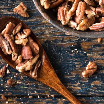 Pecans fill a wooden spoon beside a bowl, resting on a rustic wooden table.