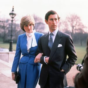 A man and woman walk arm-in-arm in formal attire, outdoors by a path and park, with a lamp post and trees in the background.