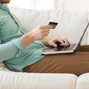 Person holding a credit card while using a laptop, seated on a white sofa with casual attire.