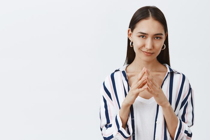 I have great evil plan. Good-looking confident and tricky woman in striped blouse, holding fingers together and smirking at camera, having some bad intention in mind, planning to commit crime