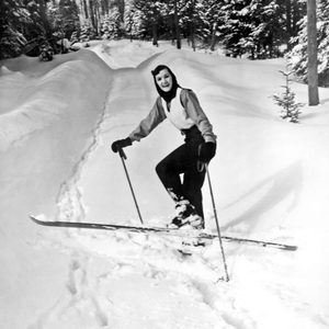 A person skis downhill on a snowy path surrounded by snow-covered trees, smiling and wearing ski gear.