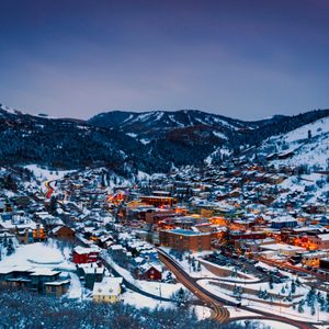 Snow-covered town illuminated at dusk, nestled in a mountainous valley with winding roads and clustered buildings surrounded by dark, forested hills.