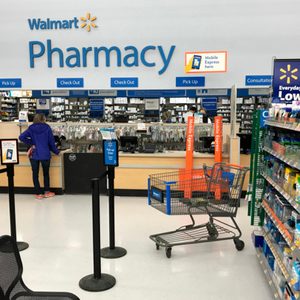 Person waits at pharmacy counter inside Walmart, surrounded by aisles of health products and a shopping cart nearby.