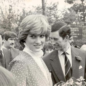 A woman smiles holding flowers, accompanied by a man in a suit. They're outdoors with people and photographers in the background.