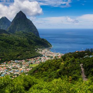Mountains rise near a coastal village, surrounded by lush greenery and a vast blue ocean under a partly cloudy sky.