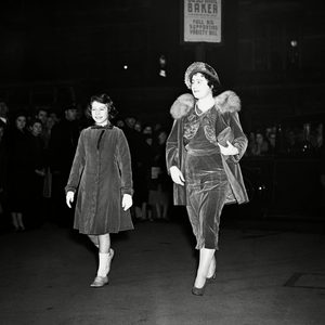 Two elegantly dressed women walk, smiling, in front of a crowd at night. A marquee reads 