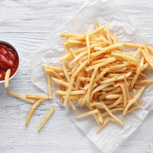 Golden French fries rest on parchment paper; a few are dipped in a small cup of red ketchup on a wooden table.