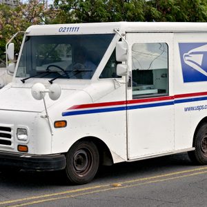 Mail truck drives on road, displaying USPS logo and website, surrounded by urban landscape with trees.