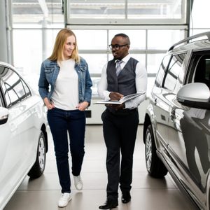 A car dealer holds a notebook while walking with a customer between two cars in a bright showroom.