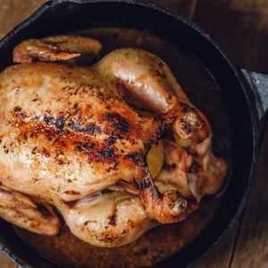 A roasted chicken rests in a cast iron skillet on a wooden table.