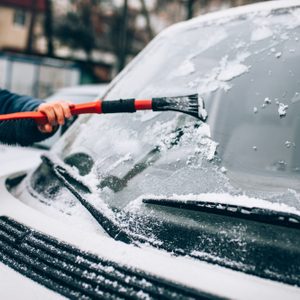 A person scrapes snow from a car windshield using a brush-scraper tool in a snowy neighborhood.