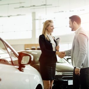Two people shake hands, smiling in a car showroom, with vehicles visible around them under bright lighting.