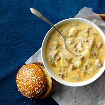 Bowl of creamy soup with visible chunks sits on parchment paper beside a sesame seed bun, resting on a blue fabric background.