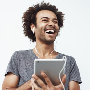 A person laughs while holding a tablet and wearing earphones, standing against a plain light background.